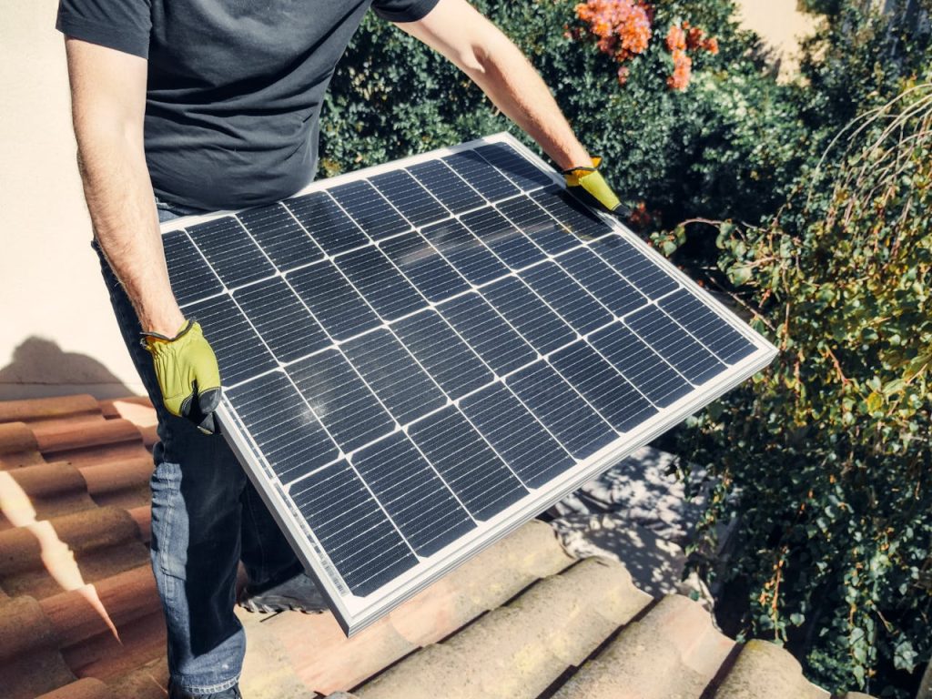 pexels-photo-9875441 A worker in gloves installs a solar panel on a tiled rooftop, promoting renewable energy.